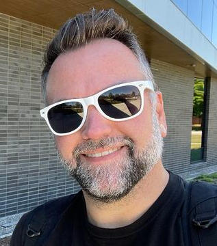 Andy wearing white sunglasses outside a building smiling at the camera.